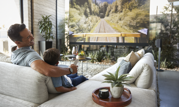 A father and son watch TV in a brightly lit living room.